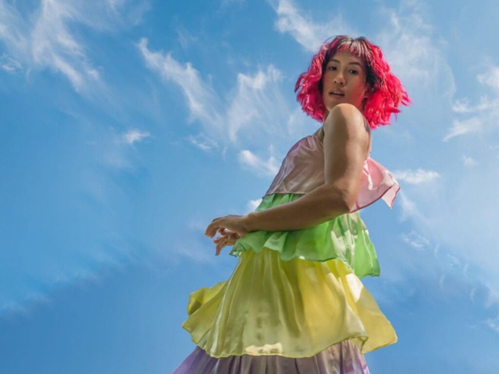Image by Sy Chounchaisit. 

Photo of Sy with a short bob haircut dyed in a vibrant hot pink shade. They're wearing a multicolor tiered dress that whimsically flows in the summer breeze. 