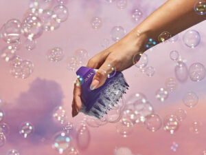 Manicured hand holding our Aura scalp massager surrounded by bubbles.
