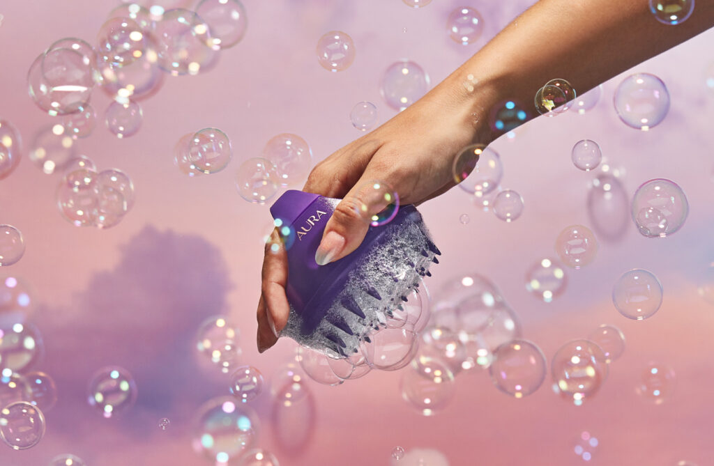 Manicured hand holding our Aura scalp massager surrounded by bubbles.