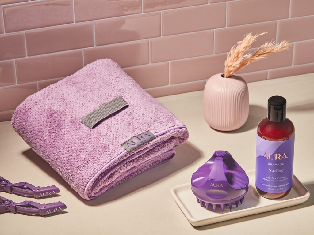 Our AURA Mirror Moments Hair Wrap Towel, Mirror Moments Hair Clips, Scalp Massager, and shampoo on a bathroom counter in front of a pink tile back splash.