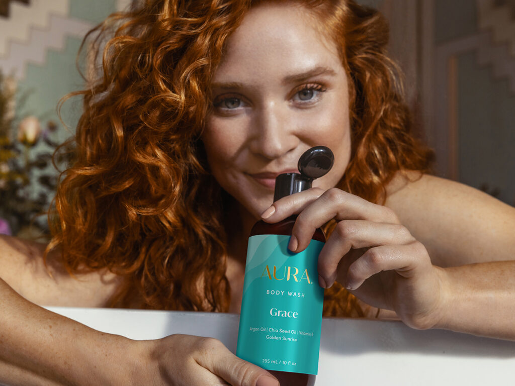 A woman with curly red hair, smiling with an open bottle of our bady wash in her hands. 