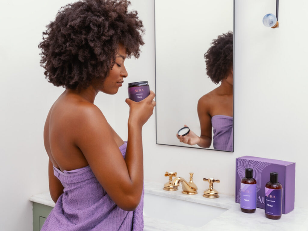 Woman of color wrapped in a purple towel smelling her personalized masque in the bathroom.