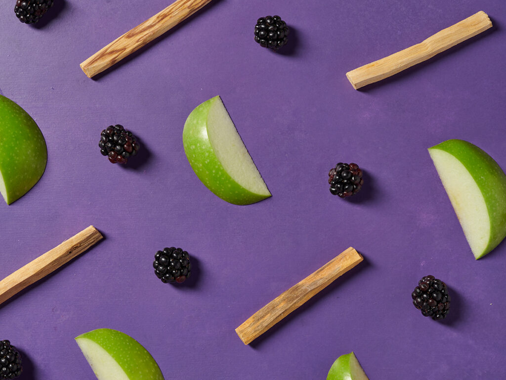 Apple slices, blackberries, and pieces of sandalwood against a purple background. 
